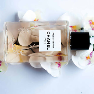 Eine elegante Flasche Chanel Parfüm umgeben von zarten weißen Orchideenblüten auf einem hellen Hintergrund. Die minimalistische Glasflasche zeigt das klassische Chanel-Label und ist künstlerisch mit Blütenblättern dekoriert.