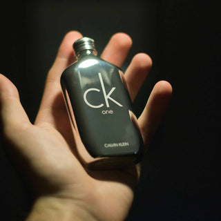 Eine Hand hält eine dunkle Glasflasche des ikonischen CK One Parfüms von Calvin Klein vor einem schwarzen Hintergrund, künstlerisch beleuchtet