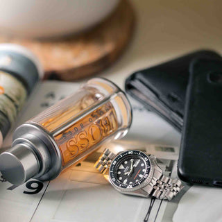 Luxuriöse Stillleben-Aufnahme mit einer Boss Parfümflasche in Goldtönen, einer silbernen Seiko Taucheruhr mit schwarzem Zifferblatt und einem schwarzen Smartphone auf einem hellen Untergrund