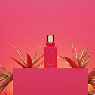 Eine rote Parfümflasche von Azalea Moscow auf einem rosa Podest, flankiert von zwei goldenen Aloe-Vera-Pflanzen vor einem rosa-orangenen Farbverlauf Hintergrund. Minimalistisches Luxus-Parfüm Produktfoto im modernen Design.