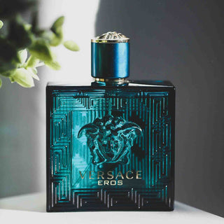 Versace Eros Parfüm in türkisblauer Glasflasche mit geometrischem Muster und Medusa-Logo auf schwarzem Hintergrund neben grüner Pflanze