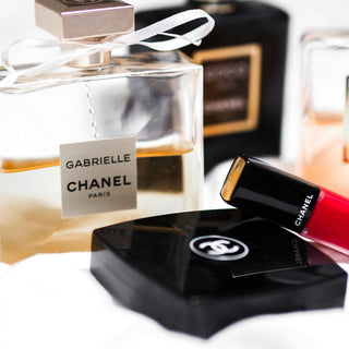 Chanel Gabrielle Parfüm - Eleganter transparenter Glasflakon mit weißer Schleife und goldenem Inhalt neben schwarzer Chanel Kosmetik und rotem Lippenstift, luxuriöse Beauty-Komposition in Nahaufnahme